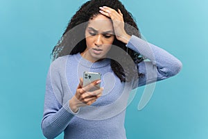 Shocked curly brunette young woman holding smartphone, looking at mobile phone screen and has disappointment face