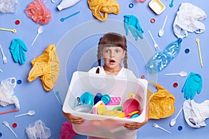 Shocked cute little girl wearing white t shirt posing against blue wall and much plastic garbage around, holding plastic toys in