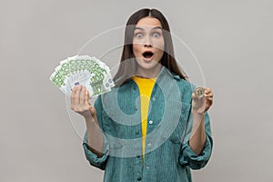Shocked dark haired woman holding golden coin of crypto currency and big fan of euro banknotes.