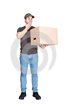 Shocked delivery man, full length portrait, holding a cardboard box while cover his mouth with hand, isolated on white. Astonished