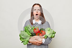 Shocked displeased brown haired young woman embraces bouquet of fresh vegetables wearing striped casual shirt isolated over gray