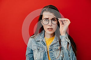 Shocked emotional young woman keeps hand on rim of spectacles, dressed in denim jacket, gazes with stupefaction
