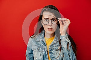 Shocked emotional young woman keeps hand on rim of spectacles, dressed in denim jacket, gazes with stupefaction photo