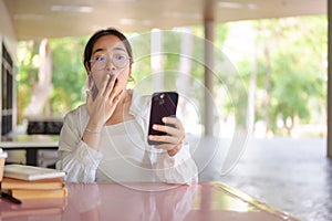 A shocked, excited Asian female college student covering her mouth while looking at her smartphone
