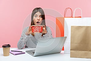 Shocked excited woman office worker holding and showing sale card looking with astonishment, sitting at workplace with laptop and
