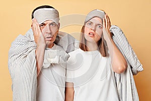 Shocked exhausted couple man and woman in sleep eye mask wrapped in blanket isolated over beige background looking at camera with