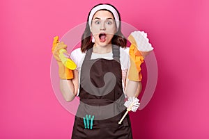 Shocked female housekeeper holds sponge and detergent spray in hands, having much work to do. Attractive woman with surprised and