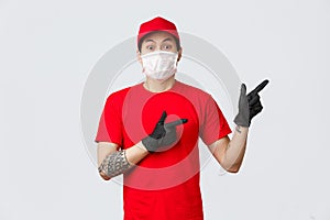 Shocked and freak out asian delivery guy in medical mask, protective gloves and red uniform pointing fingers right