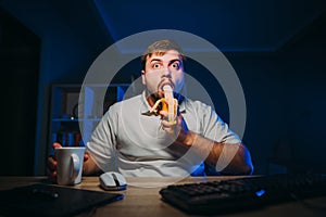 Shocked freelancer man with cup in hands works at night on computer in dark room and eats banana with surprised face looks at