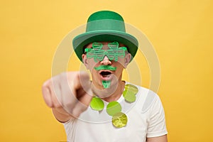 Shocked frighten scared man wearing green leprechaun hat and shamrock glasses posing isolated over yellow background looking with