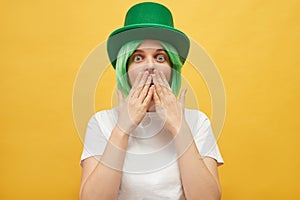 Shocked frighten woman with green hair wearing leprechaun hat posing isolated over yellow background looking at scared eyes sees