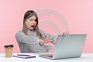 Shocked frightened brunette woman office worker raising hands in stop gesture looking at laptop screen, unpleasant video call,