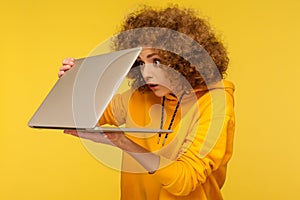 Shocked frightened woman with curly hair in urban style hoody looking furtively at half closed laptop screen