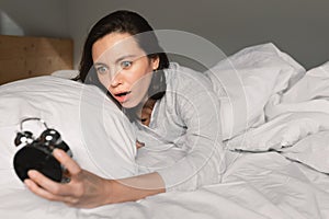 Shocked frightened young european woman lies on white comfortable bed, wake up, looks at alarm clock