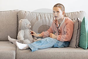 Shocked funny caucasian little girl playing doctor with toy bear, listens to breath with stethoscope