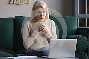 Shocked grey haired mature woman reading unexpected news on laptop