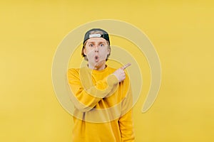 Shocked guy in a cap and yellow sweatshirt and cap shows his finger to the side and looks at the camera with a surprised face