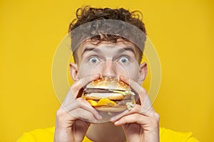 Shocked guy eats a big burger and is surprised on a yellow background, a man bites fast food, close-up