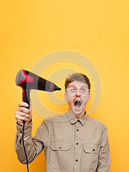 Shocked guy in glasses and mustache uses hairdryer on yellow background, looks up with surprised face on copy space. Emotional