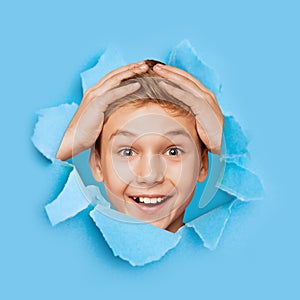 Shocked happy excited european teen boy holding his hands behind head looking through hole on blue paper