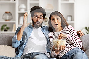 Shocked hindu couple sitting on couch at home, watching triller
