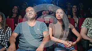 Shocked Hispanic man and woman watching film with amazement face expression surrounded by spectators