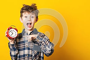 Shocked kid boy holding clock alarm, copy space. Kid isolated over yellow background. Time for school. Little student overslept. T