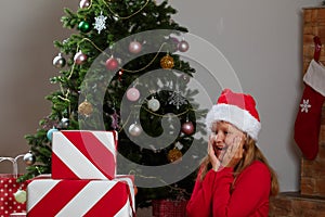 A shocked little girl in a Santa Claus hat looks at a stack of boxes with gifts on the background of a Christmas tree