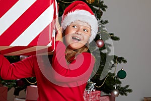 A shocked little girl wearing a Santa Claus hat holds a large Christmas box with a gift.