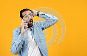 Shocked man in a blue denim shirt talking on a cellphone, holding his head in disbelief or receiving