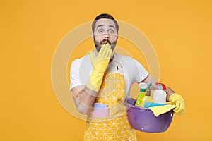 Shocked man househusband in apron rubber gloves hold basin with detergent bottles washing cleansers doing housework