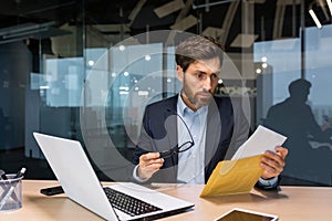 A shocked man in the office, holding an envelope with a letter with bad news, bill, credit