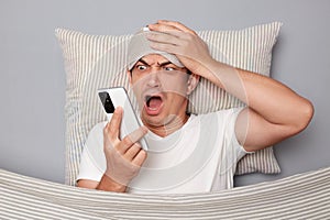 Shocked man with resting on white pillow under blanket wearing t-shirt isolated over grey background waking up looking at