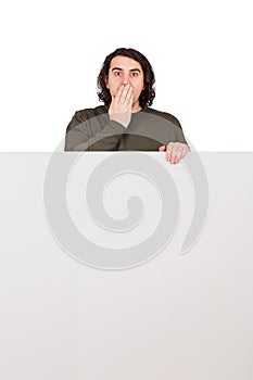 Shocked man, staring to camera with big eyes, covers his mouth with hand, stands behind a blank banner for announcements. Empty