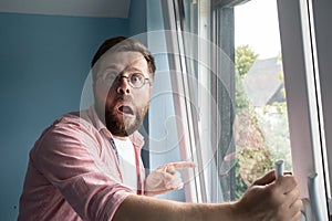 Shocked man with a strange expression on face stands near an open window and points finger into the courtyard in