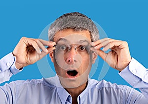 Shocked mature man looking at camera through eyeglasses, blue background