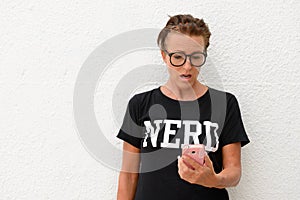 Shocked mature nerd woman wearing big eyeglasses and standing against white background outdoors while using mobile phone