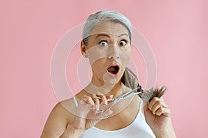 Shocked middle aged Asian lady cuts split ends of grey hair on pink background