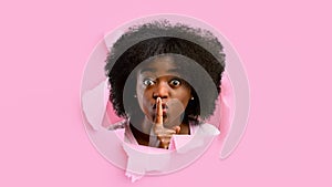 Shocked millennial black female looks into hole in pink paper, shows finger near lips, sign of silence