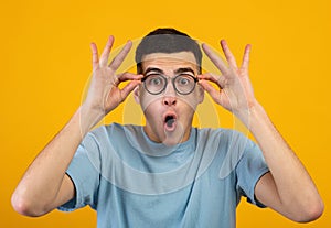 Shocked millennial guy touching his glasses, shouting WOW or OMG on orange studio background