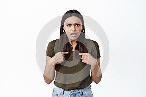 Shocked and offended young woman pointing at herself with displeased face, being insulted, feeling bad, standing against photo
