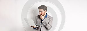 Shocked office worker in suit looking worried at laptop screen, having trouble at work, standing on white background