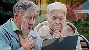 Shocked old married couple looking at laptop screen sitting in autumn park upset elderly man and woman reading bad news