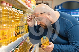 Shocked overweight man raising his glasses to his forehead and looking on price of vegetable oil in supermarket. Amazed