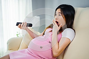 Shocked pregnant woman watching tv with remote control on sofa in living room