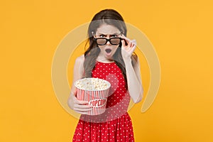 Shocked puzzled young brunette woman girl in red summer dress, 3d glasses isolated on yellow wall background. People