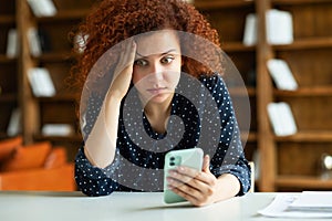 Shocked red-haired young woman sitting at the desk, holding smartphone
