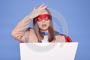 Shocked regretful brown haired woman wearing superhero costume and mask posing isolated over blue background making facepalm