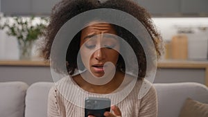 Shocked sad girl African American woman on sofa hold smartphone looking at mobile phone screen worried read bad news