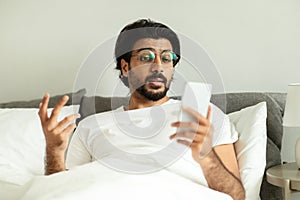 Shocked sad surprised millennial middle eastern guy in glasses, reads message on phone, lies on bed